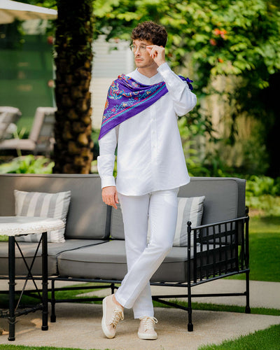 Man in a white linen mandarin collar shirt and white trousers, with a purple printed silk scarf draped over one shoulder