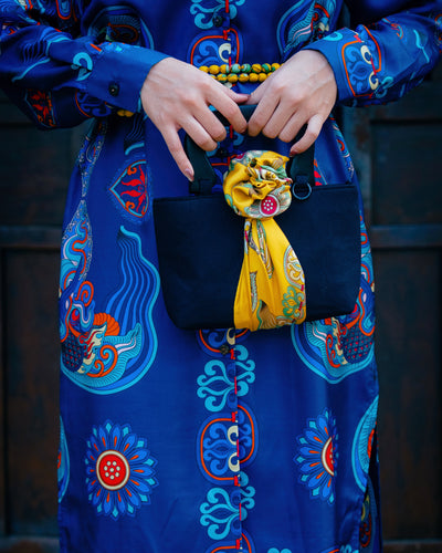 Yellow Dragon Of Grace silk scarf tied as a rosette on a black handbag handle over a blue printed dress