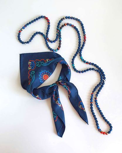 Navy blue silk scarf gathered through a ring with long pointed tails, beside a beaded necklace